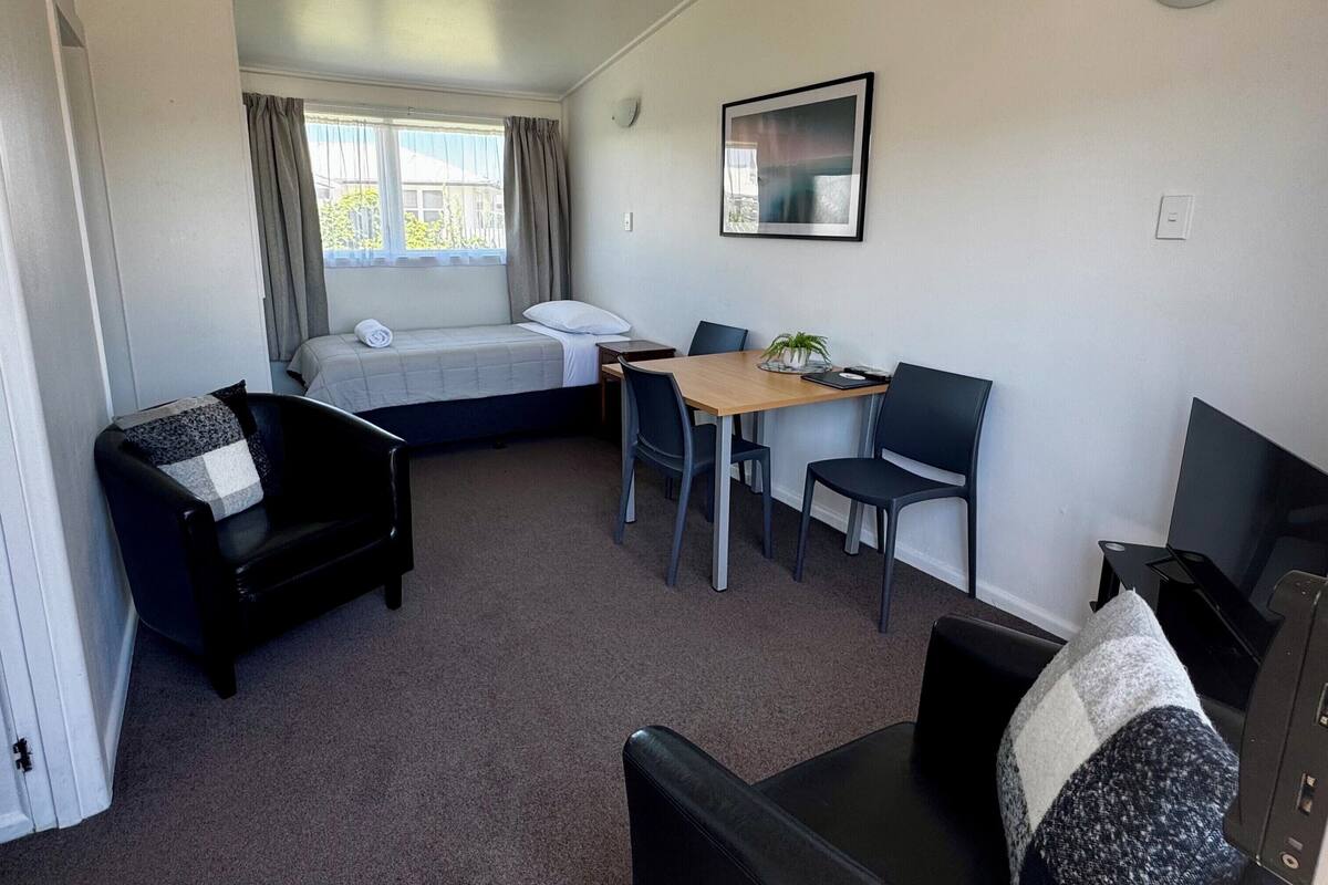 A compact room with a bed, a desk with chairs, a sofa, and a window with curtains.