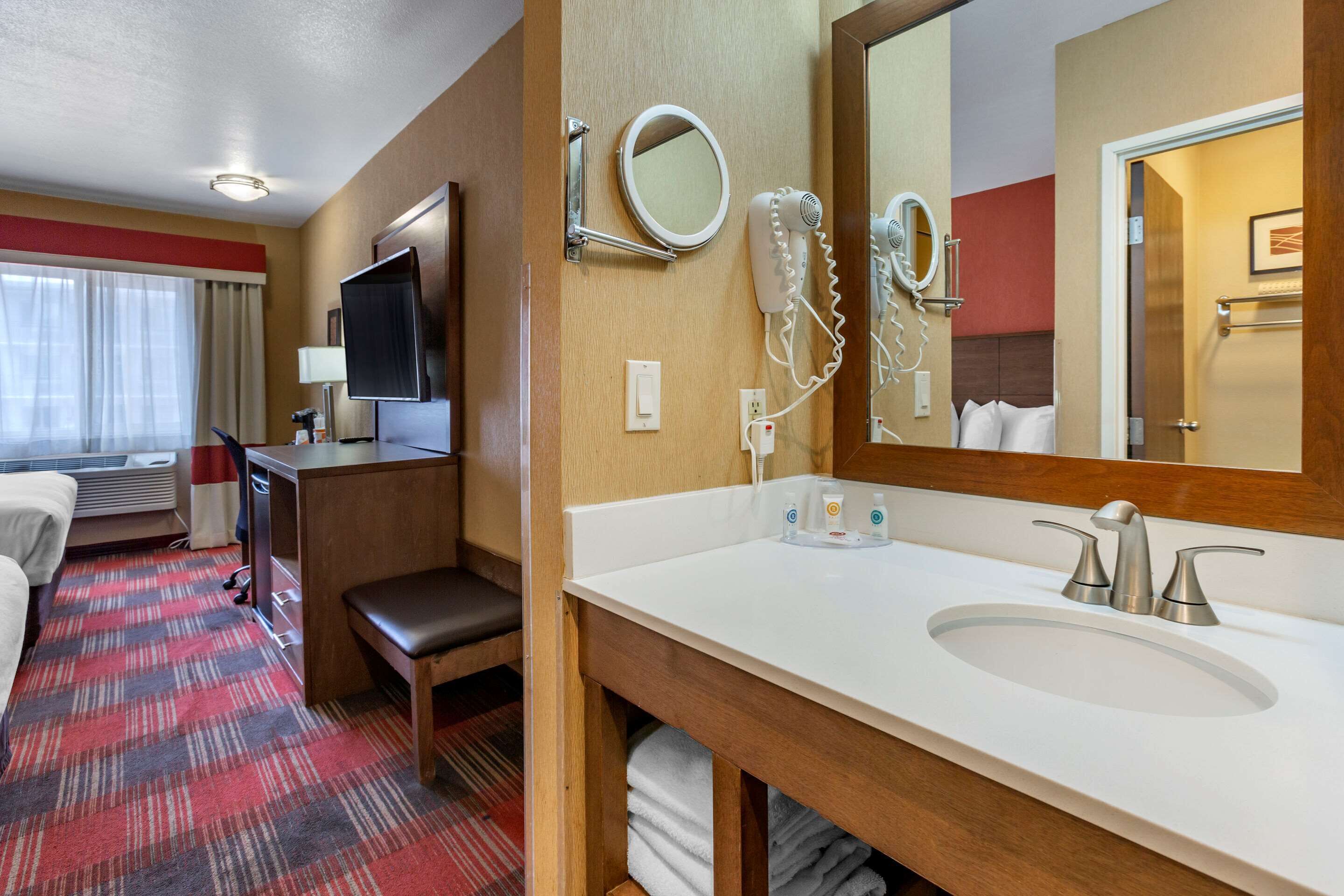 Habitación de hotel con cama, escritorio, televisor, baño con lavamanos y espejo, y piso cubierto de alfombra roja.