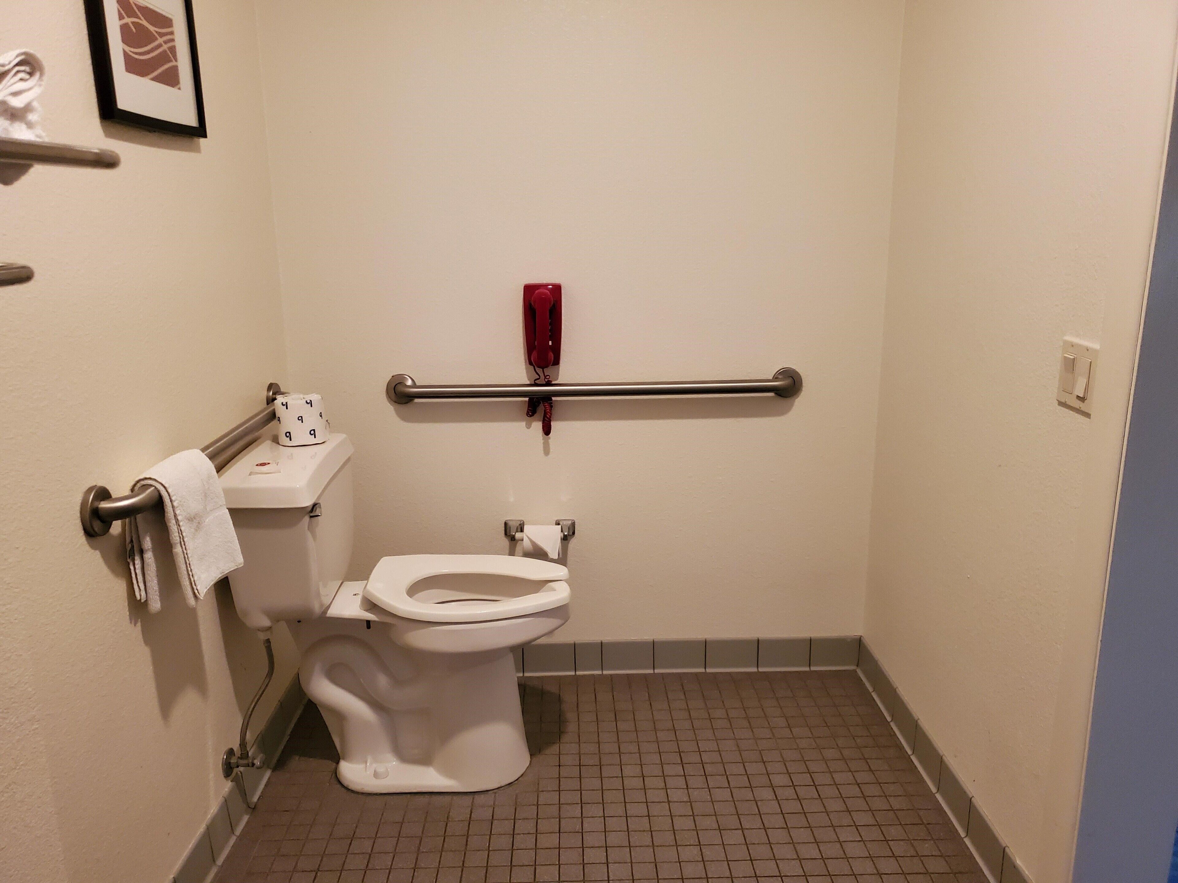 Un baño pequeño y estrecho con inodoro blanco, barra de apoyo, teléfono rojo de emergencia y un cuadro en la pared.