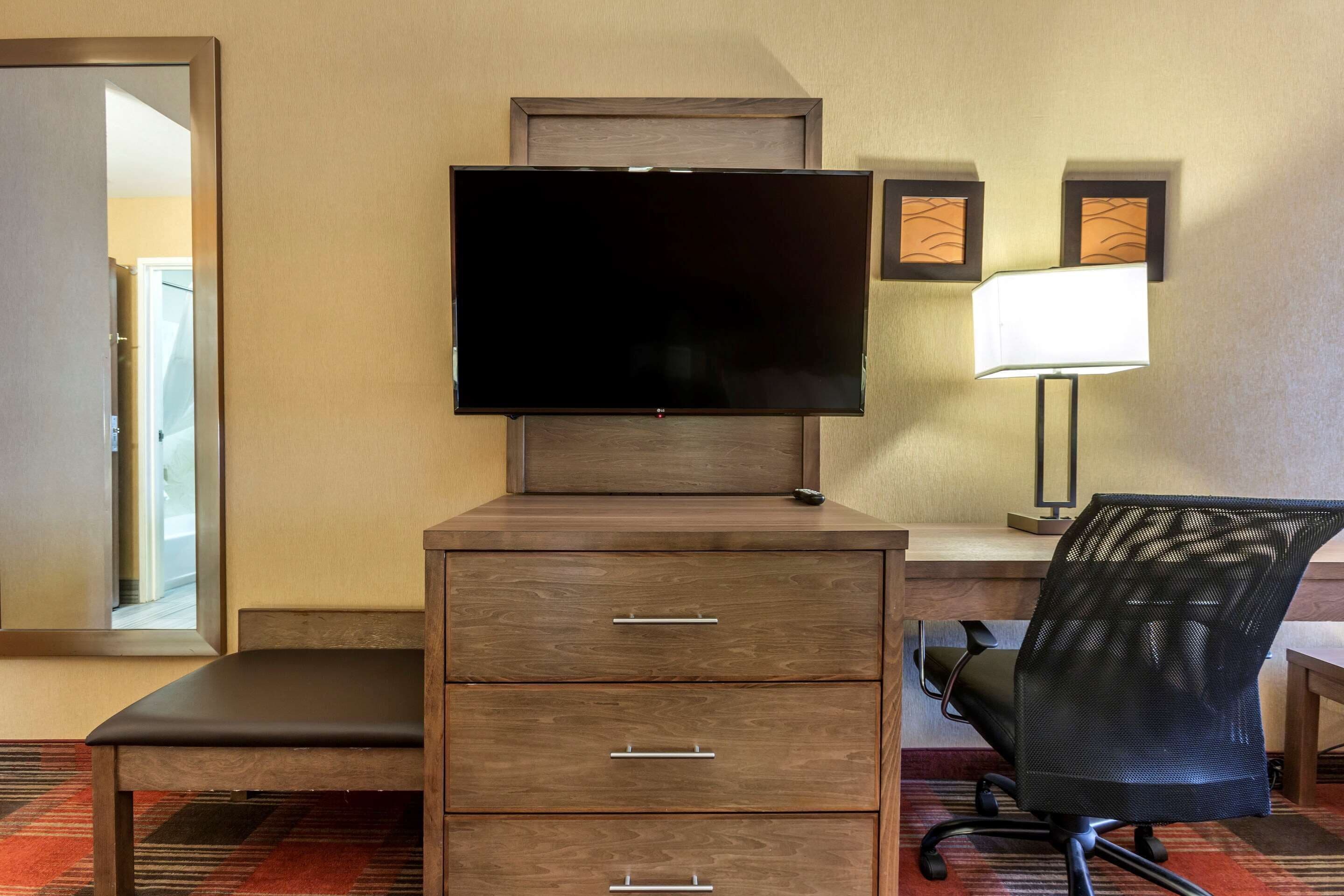 A hotel room with a TV on a wooden cabinet, a desk with a chair, and a lamp on the wall.