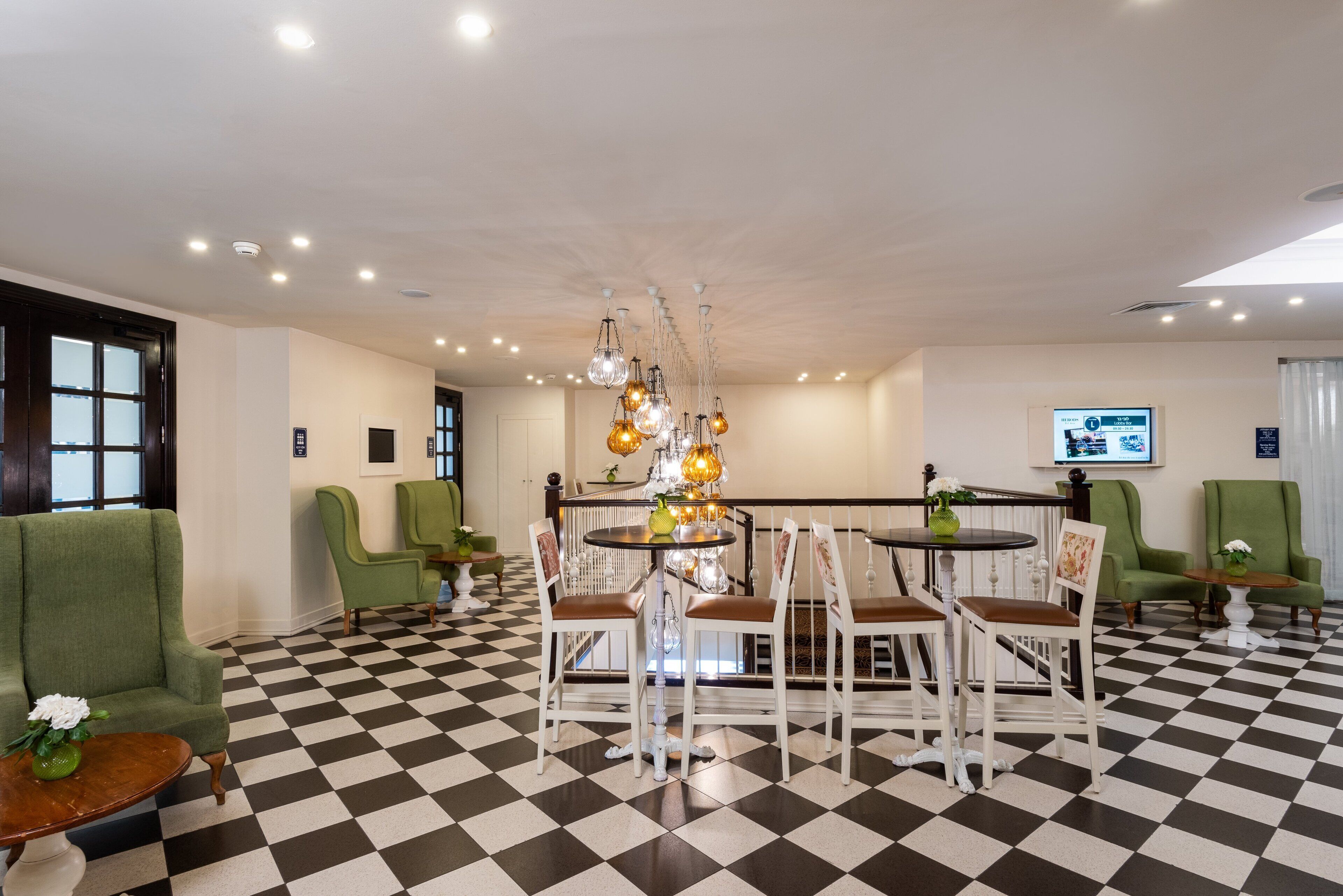 A hotel lobby with a checkered floor, green armchairs, a central table with chairs, and a chandelier.