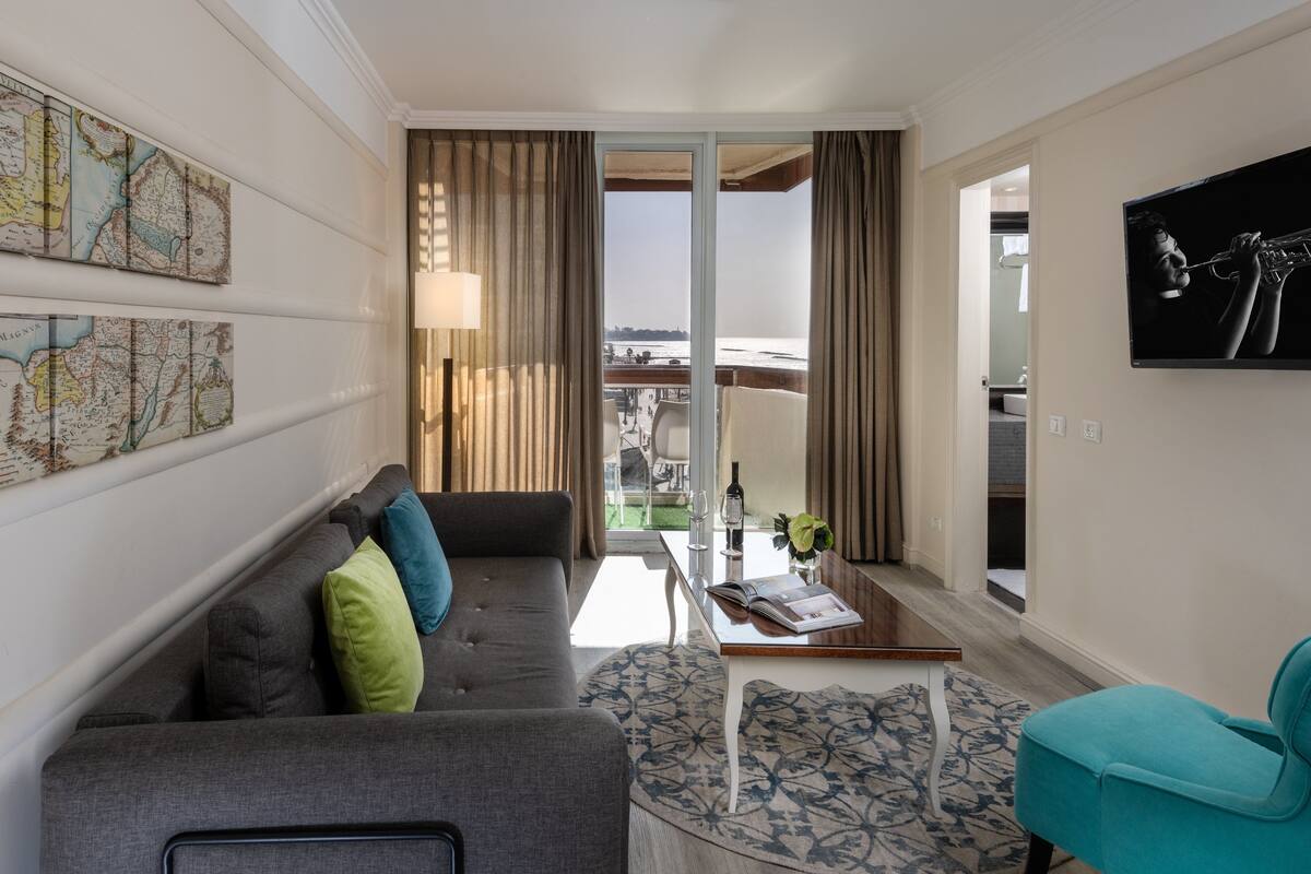 A modern living room with a grey sofa, teal armchair, and a coffee table. A large window offers a view of the outdoors.