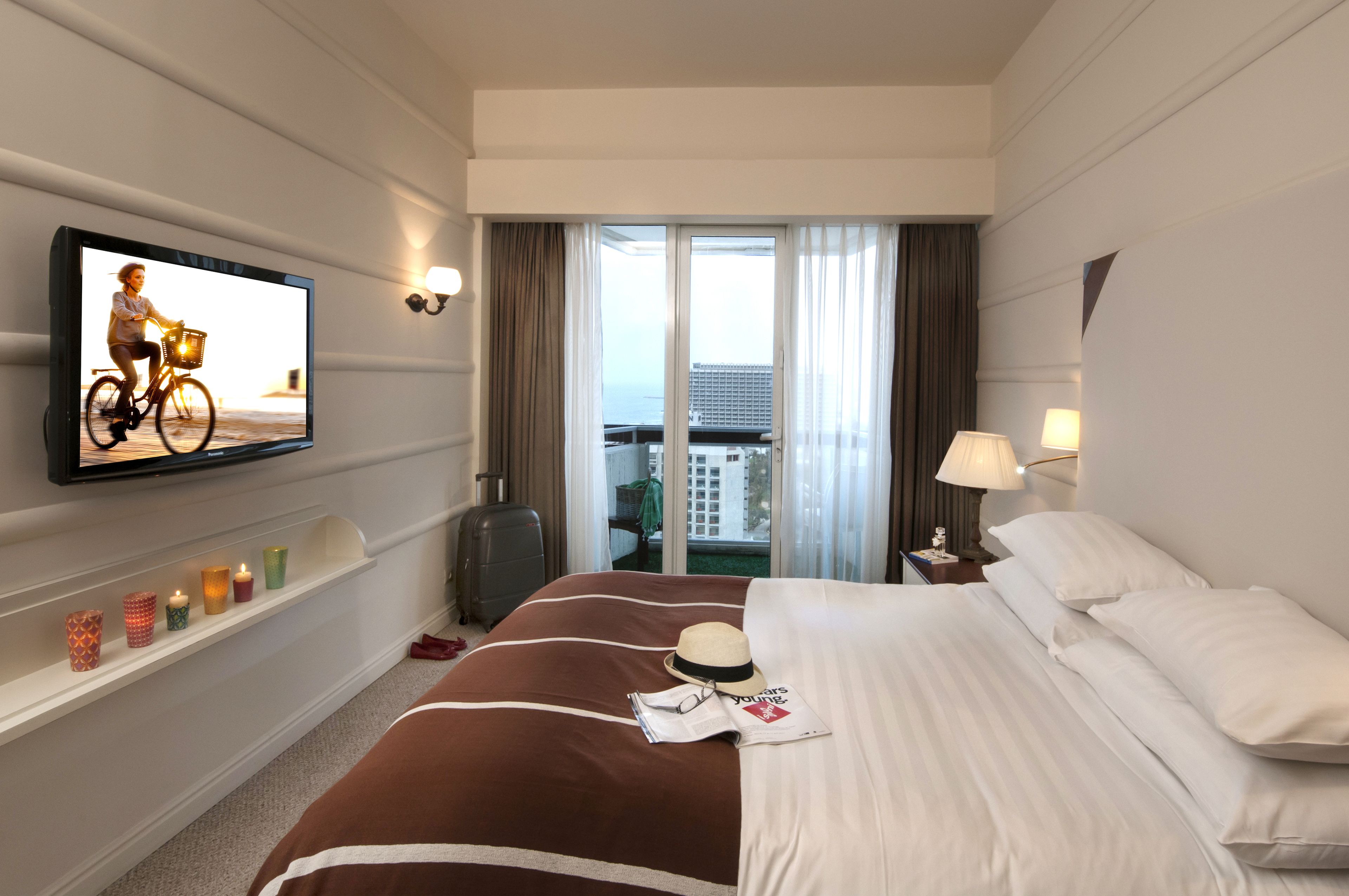 A hotel room with a bed, a television, a suitcase, a nightstand, and a view of the city through the window.