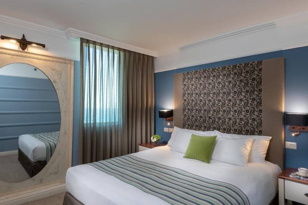 A hotel room with a large bed, a mirror, a nightstand with a lamp, and a patterned wall.