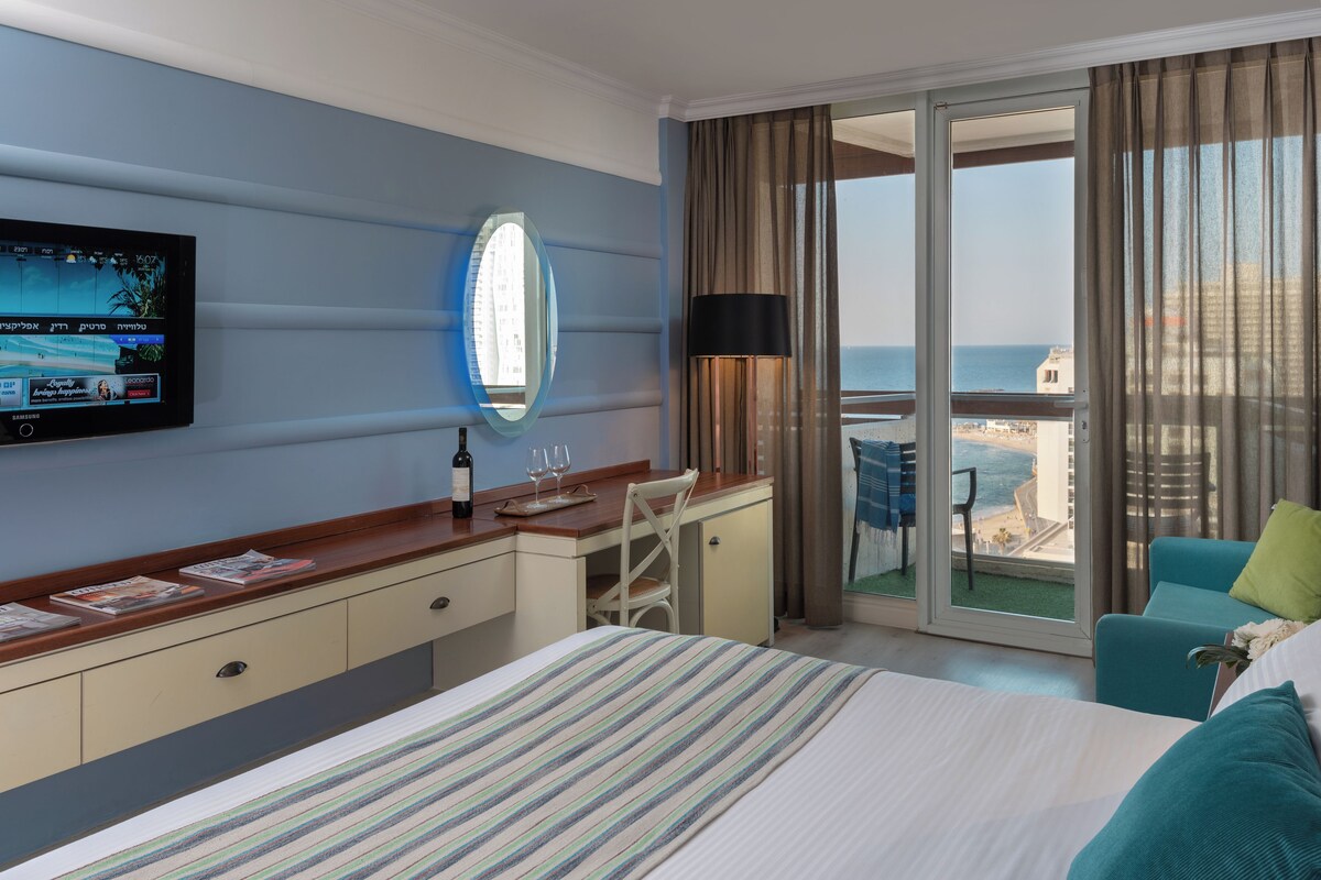 Habitación de hotel con cama, escritorio, televisor, espejo y balcón con vista al mar.