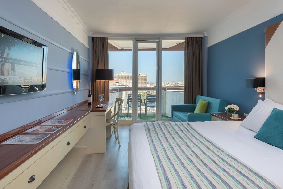 Habitación de hotel con cama, escritorio, silla, televisor y balcón con vista a la ciudad.
