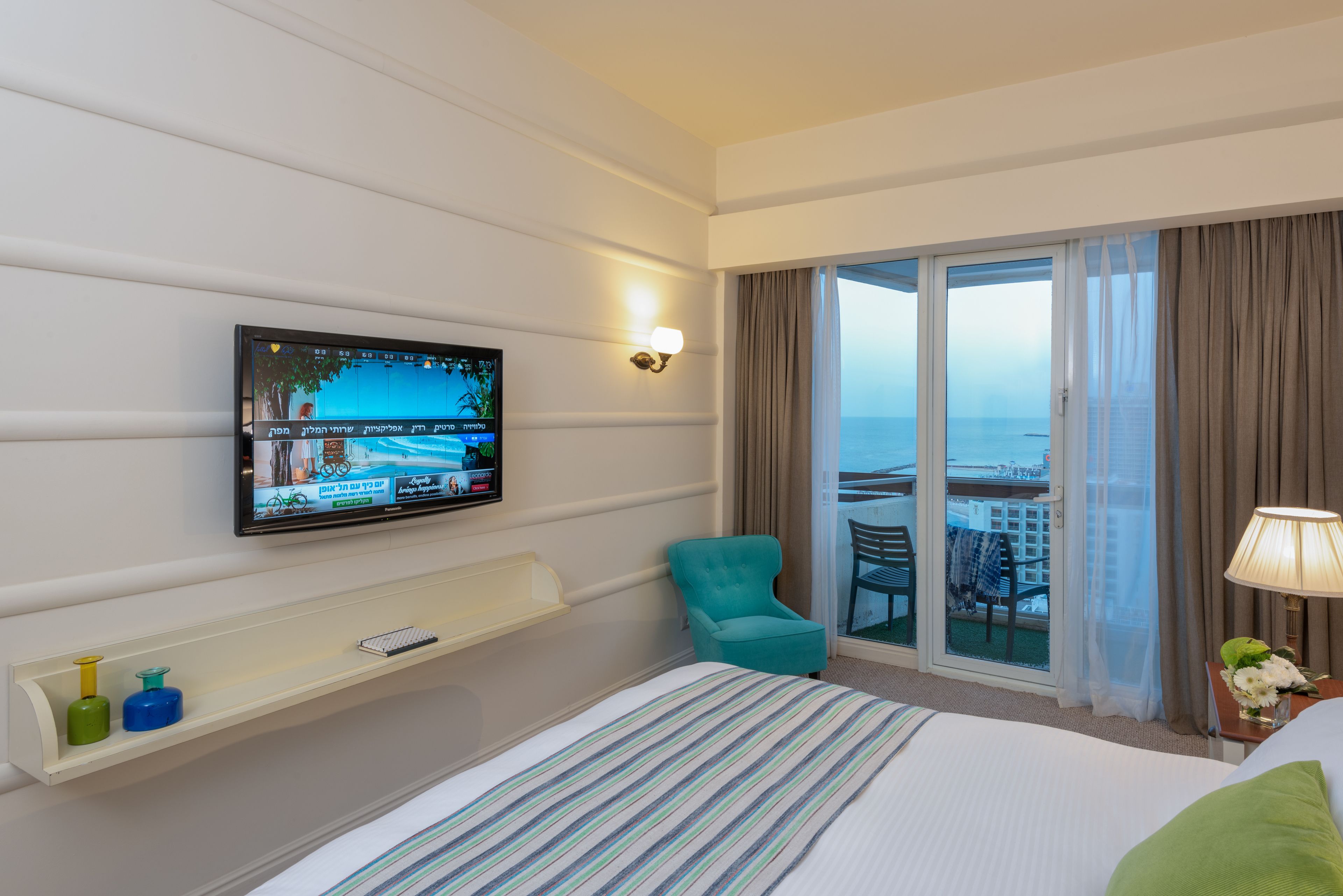Habitación de hotel con cama, televisor montado en la pared, sillón verde azulado, mesita con lámpara y vista al mar a través de una puerta corrediza de vidrio.
