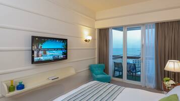 Habitación de hotel con cama, televisor montado en la pared, sillón verde azulado, mesita con lámpara y vista al mar a través de una puerta corrediza de vidrio.