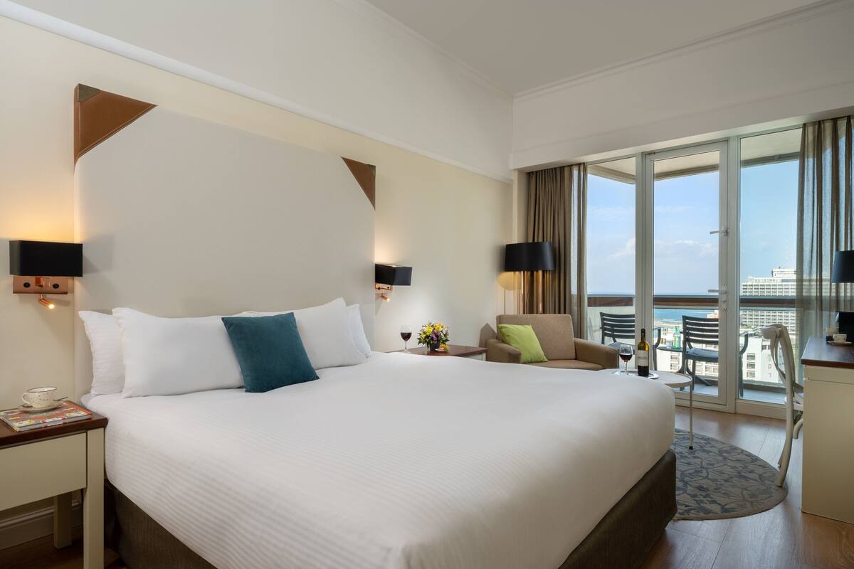 A hotel room with a large bed, a desk, a chair, a small table, and a view of the city.