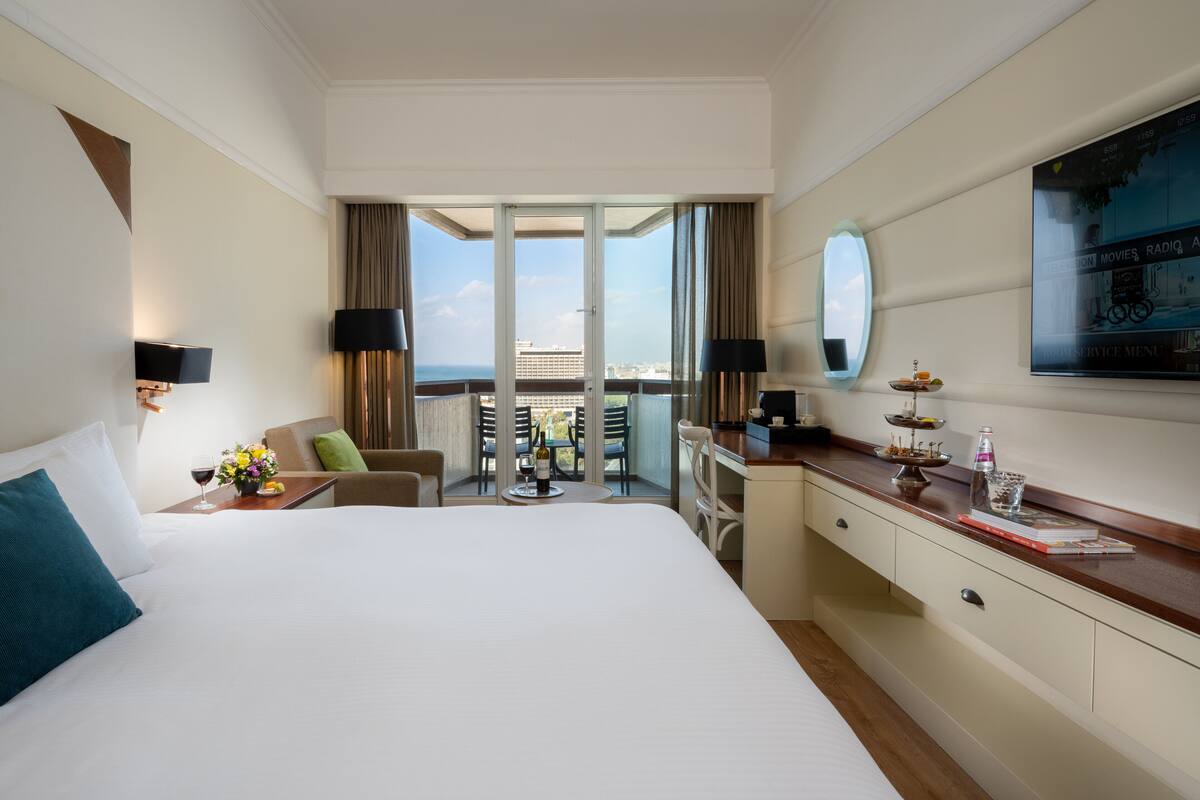 A hotel room with a large bed, a desk, a chair, a balcony with a view, and a TV.