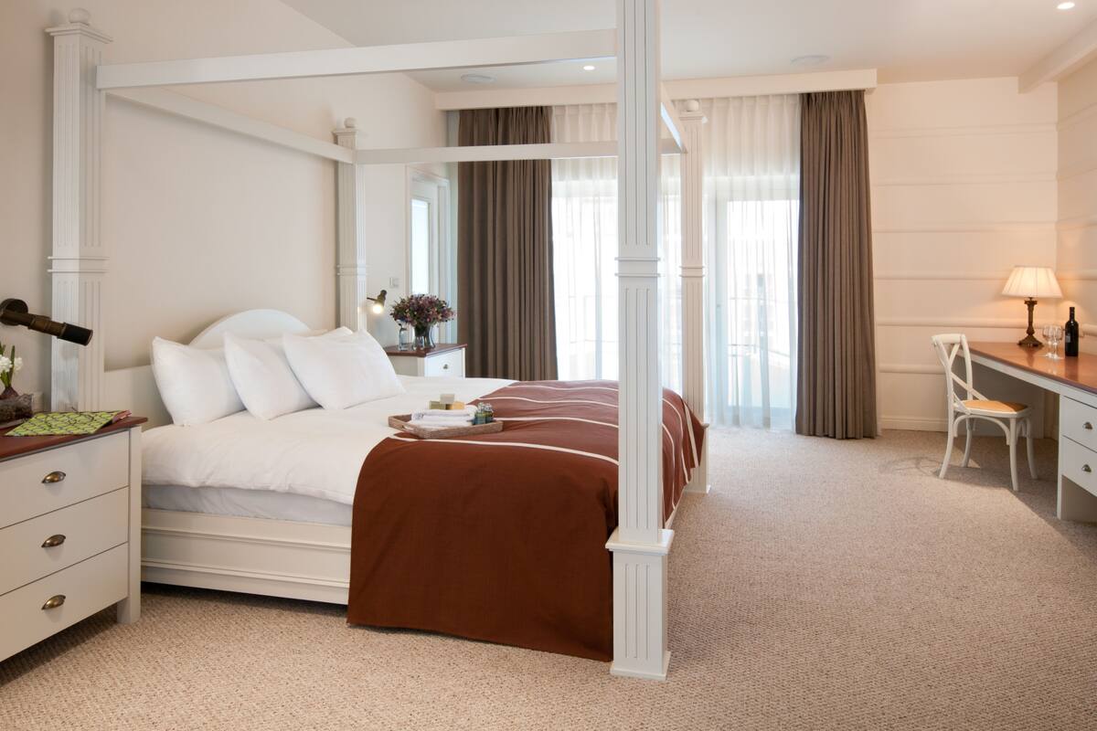A four-poster bed with white and brown bedding, a desk with a lamp, and a chair in a hotel room.