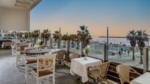 Una terraza con mesas y sillas, con vista a una playa con palmeras y gente.
