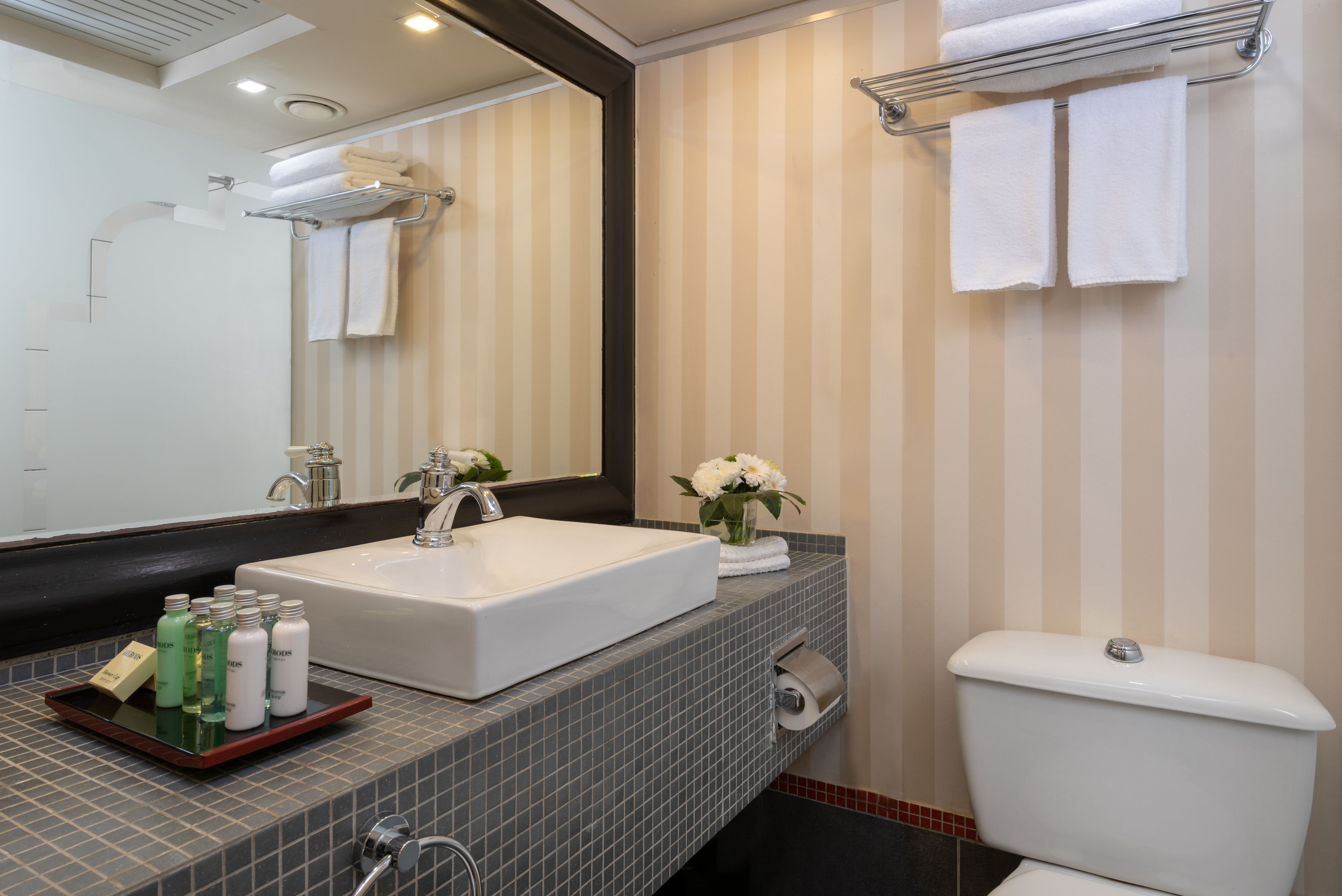 Um banheiro moderno com um espelho grande, uma pia branca e um vaso sanitário. Há toalhas penduradas em um porta-toalhas e um pequeno vaso com flores no balcão da pia.