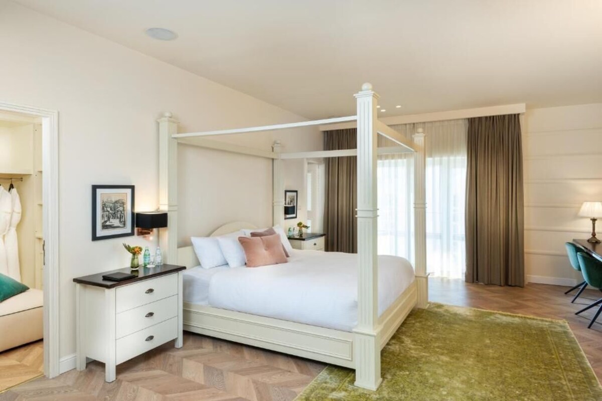 Una cama con dosel de estructura alta con acabado blanco, una mesita de noche con cajones, una silla verde y una alfombra verde.