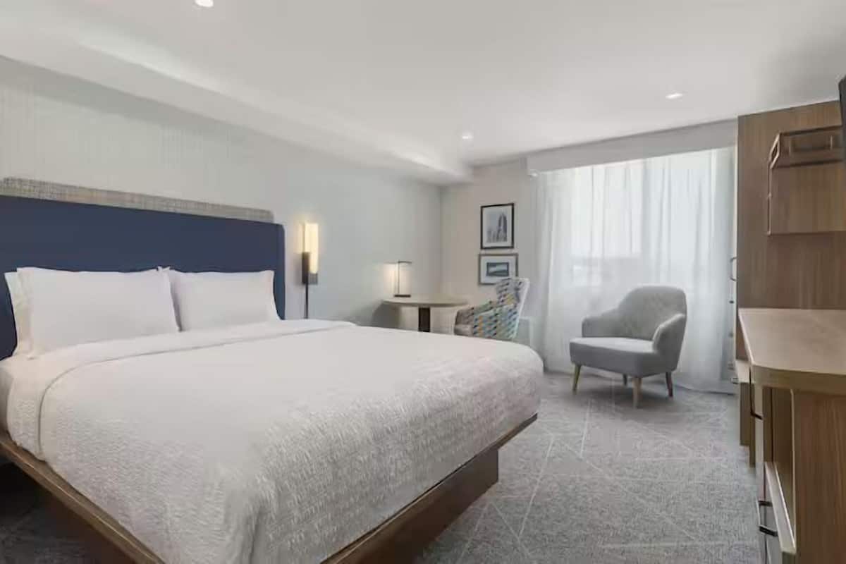 Ein Hotelzimmer mit einem großen Bett, Nachttischen, einem Schreibtisch, einem Sessel und Blick ins Freie.