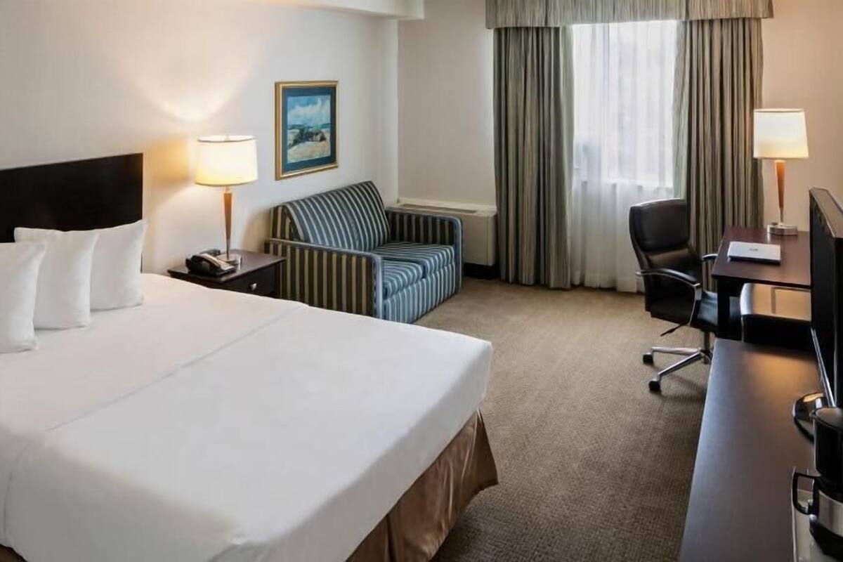 Ein Hotelzimmer mit einem großen Bett, einem Schreibtisch mit Stuhl, einem gestreiften Sessel, einem Bild an der Wand und einem Fenster mit Vorhängen.