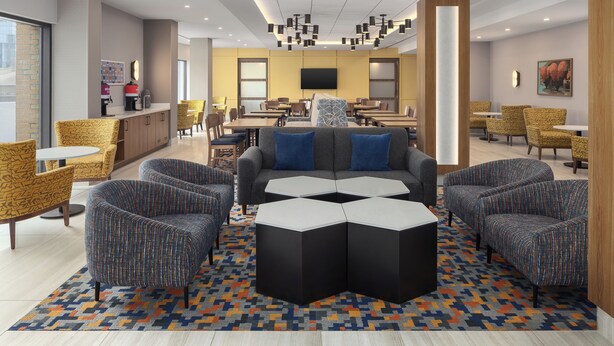 Een moderne hotellobby met een centraal zithoek, een koffiehoek en een grote flatscreen televisie aan de muur.