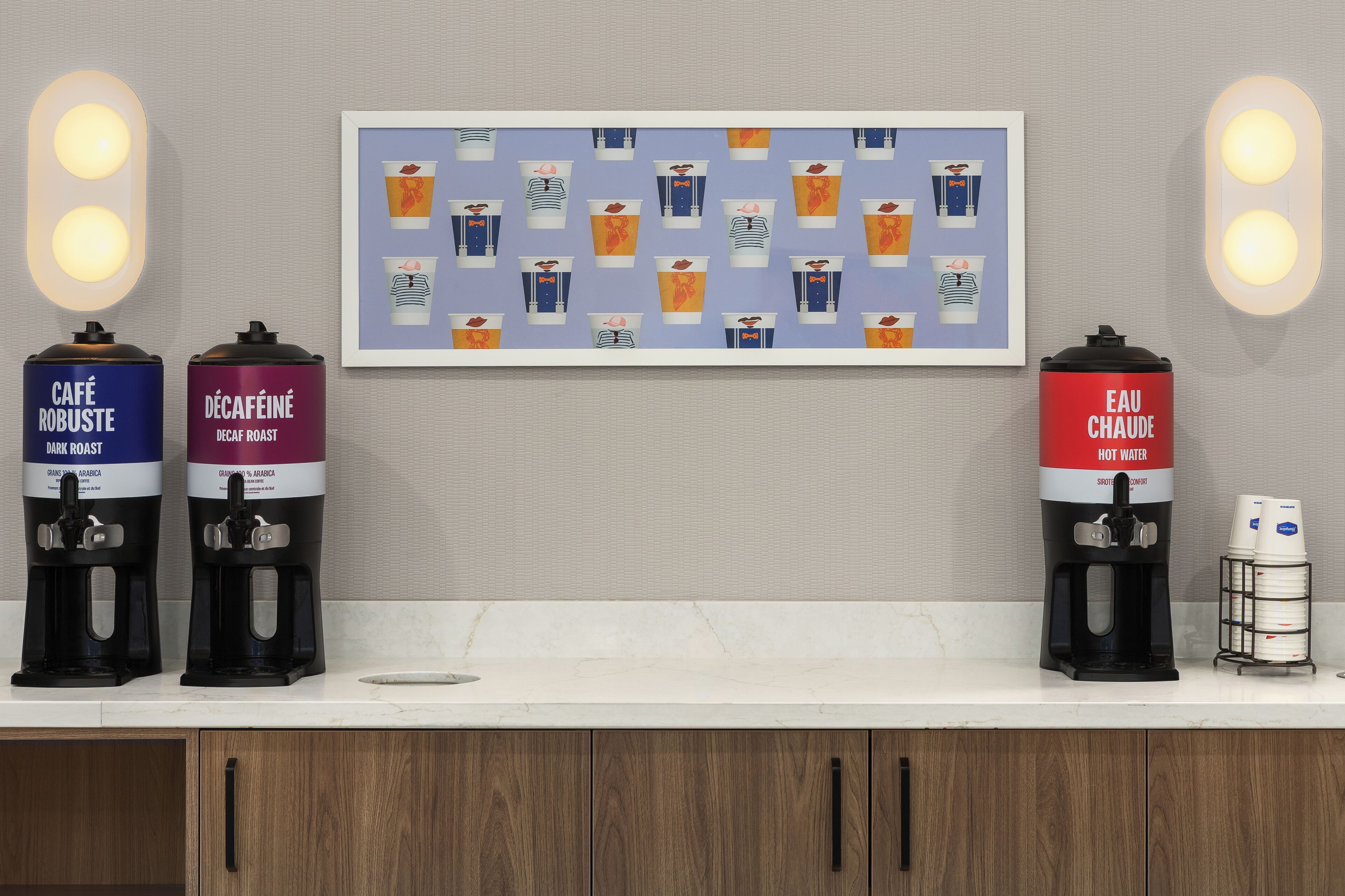 Una estación de café con dispensadores etiquetados para café y agua caliente, una obra de arte enmarcada y una lámpara fijada a la pared.