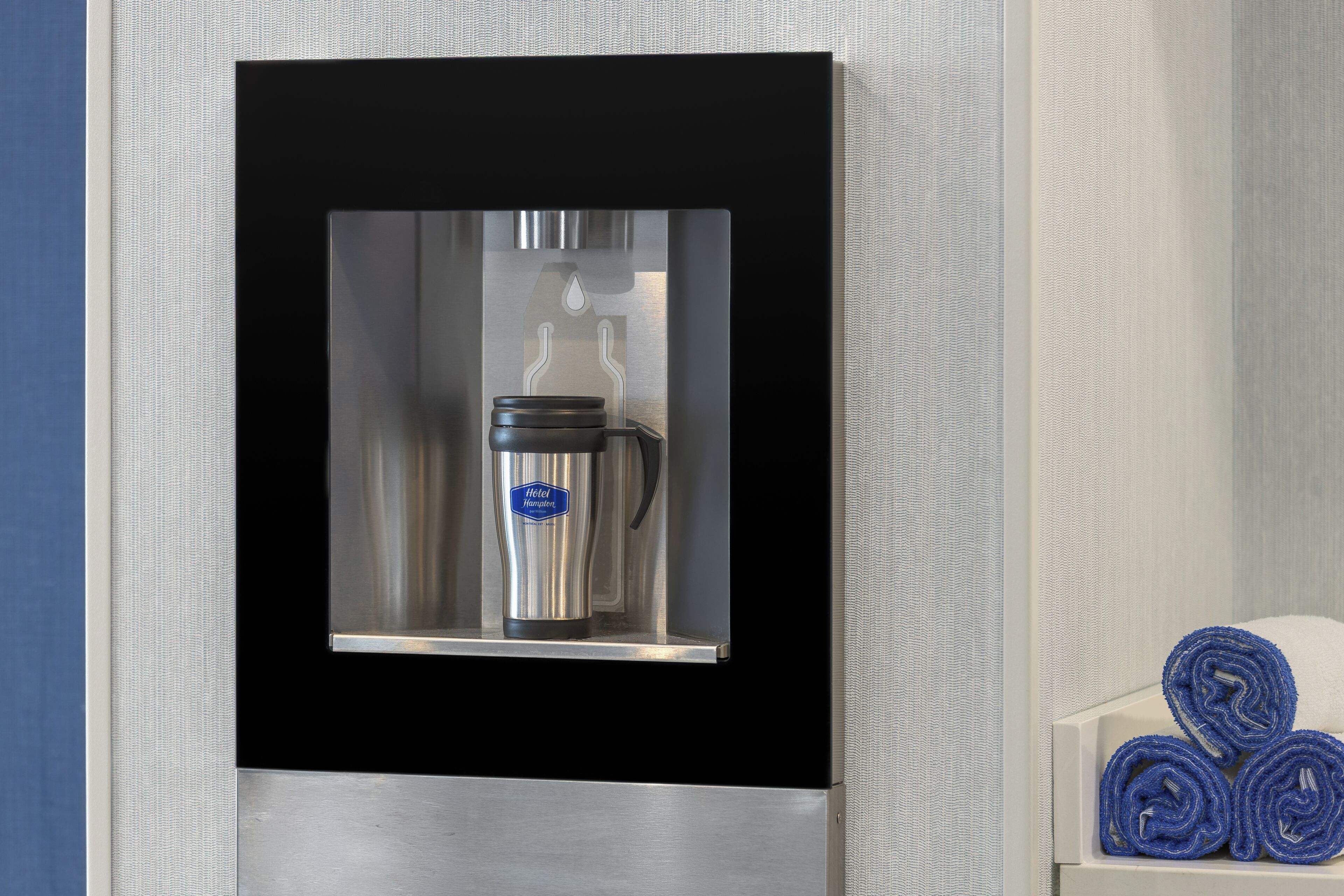 Uma máquina de café fixada na parede, com um jarro de aço inoxidável e uma etiqueta azul, ao lado de um porta-toalhas branco com toalhas enroladas.