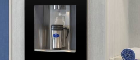 Uma máquina de café fixada na parede, com um jarro de aço inoxidável e uma etiqueta azul, ao lado de um porta-toalhas branco com toalhas enroladas.