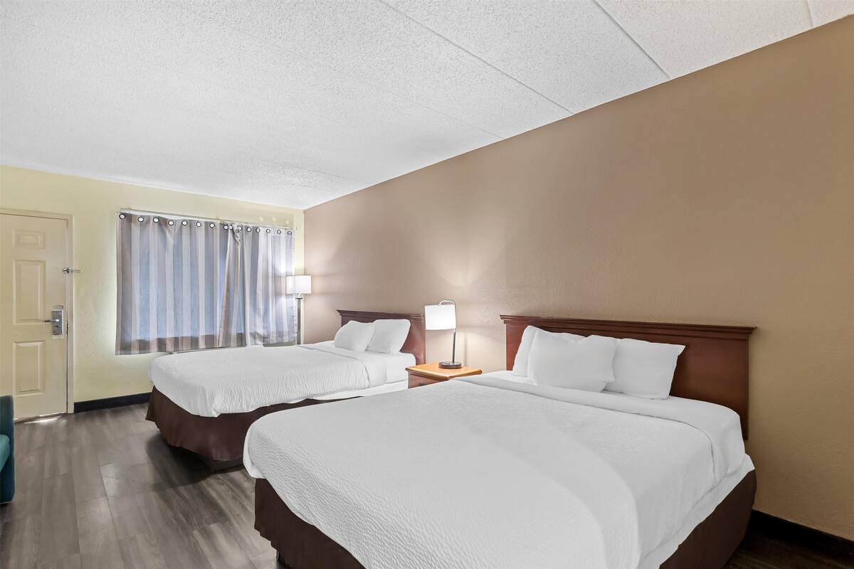 A hotel room with two beds, a door, a window with curtains, and a nightstand with a lamp.