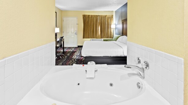 A hotel room with a large white bathtub, a bed with white linens, and a door with a curtain.