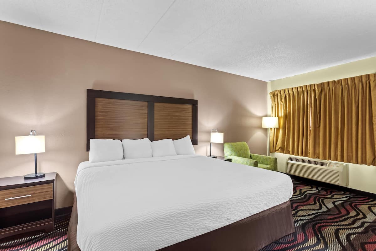 A hotel room with a large bed, two bedside lamps, a green armchair, a wall-mounted air conditioner, and a patterned carpet.