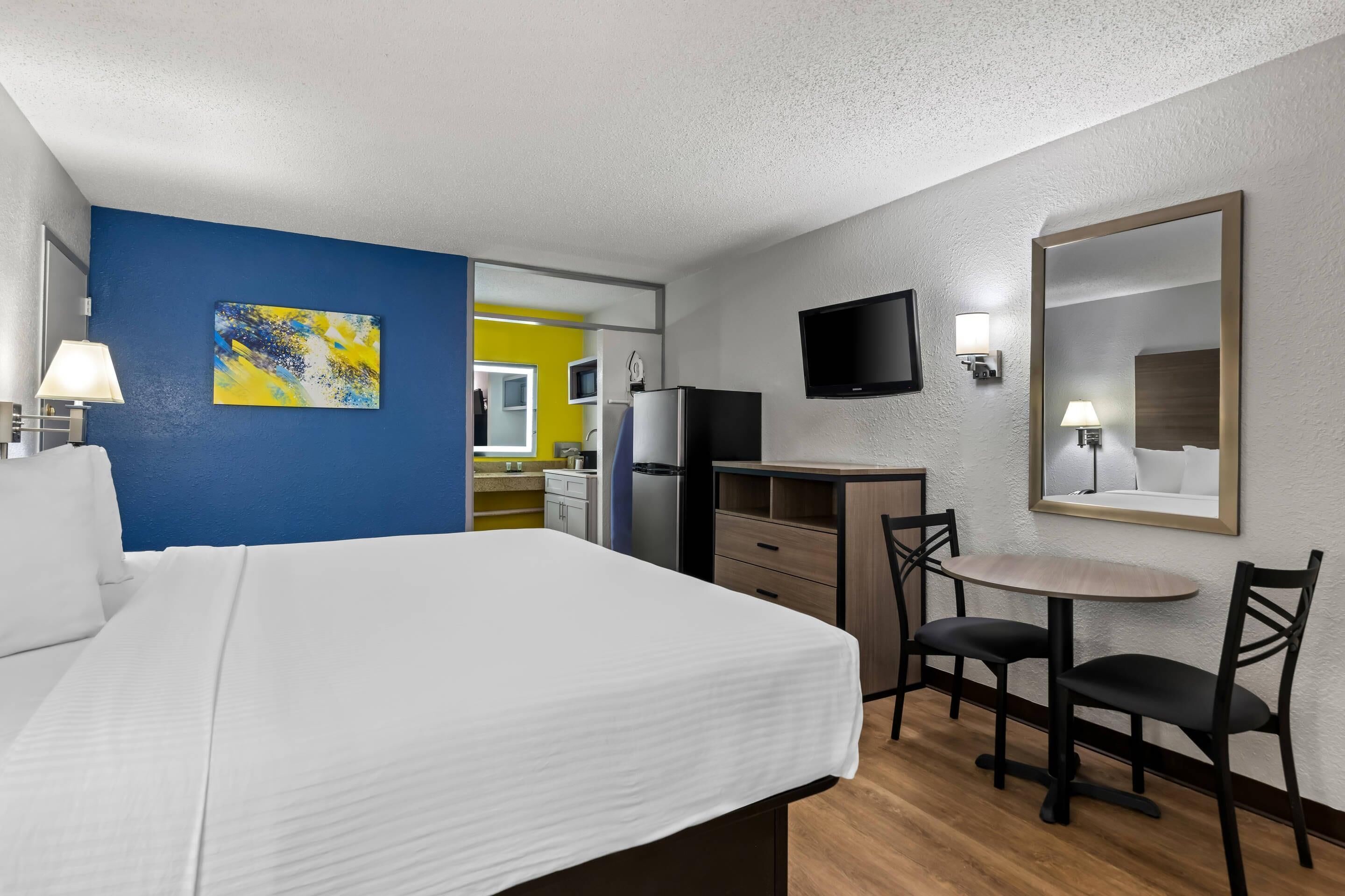 A hotel room with a bed, a desk with a chair, a TV, and a painting on the wall.