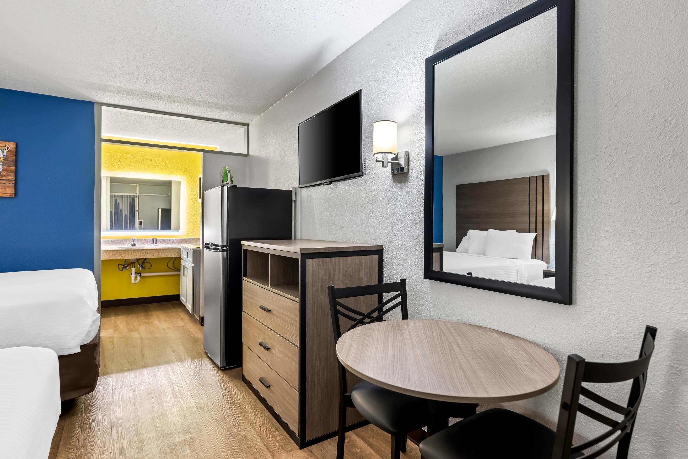 A compact hotel room with a bed, a small kitchenette, a dining table with chairs, a TV mounted on the wall, and a large mirror.