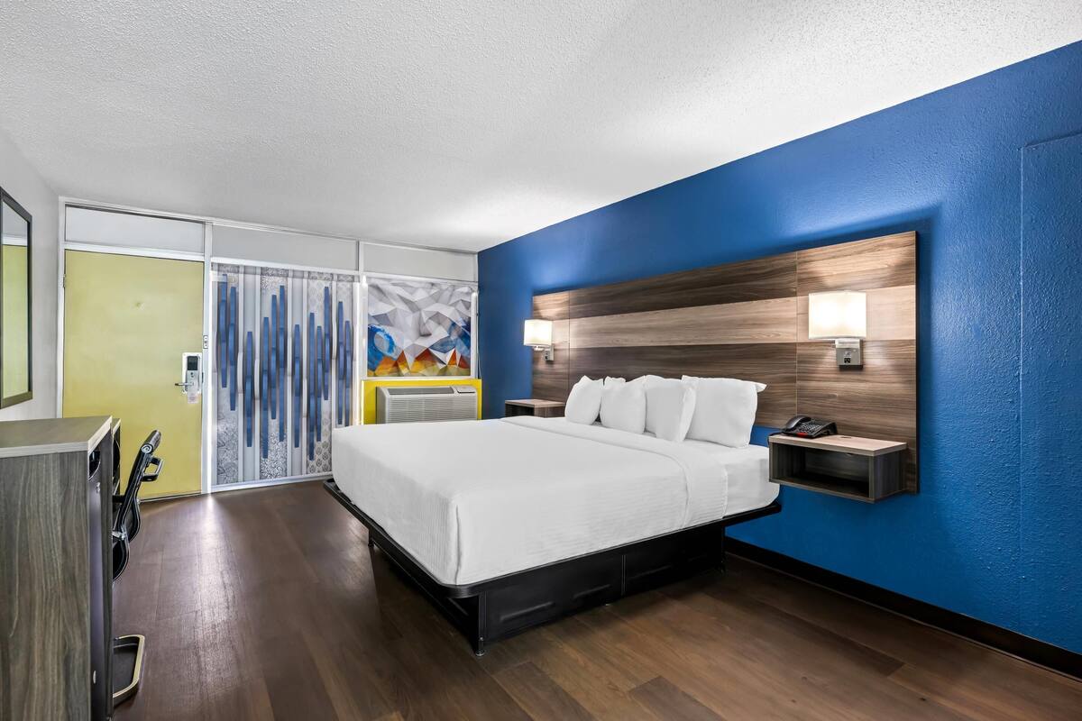 Habitación de hotel con una cama grande, un cabecero de madera, una mesita de noche con lámpara y vistas a un mural colorido a través de una puerta corredera.