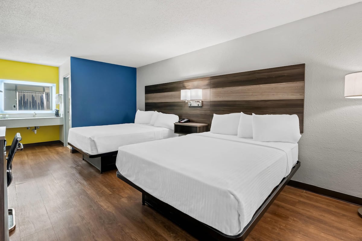 Habitación de hotel con dos camas, cabecera de madera, mesita de noche pequeña con lámpara y ventana con cortinas.