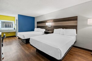 Habitación de hotel con dos camas, cabecera de madera, mesita de noche pequeña con lámpara y ventana con cortinas.
