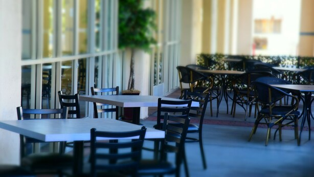 An outdoor cafe with tables and chairs arranged in rows, large windows, and a potted plant. - Ramada by Wyndham San Diego National City (National City)