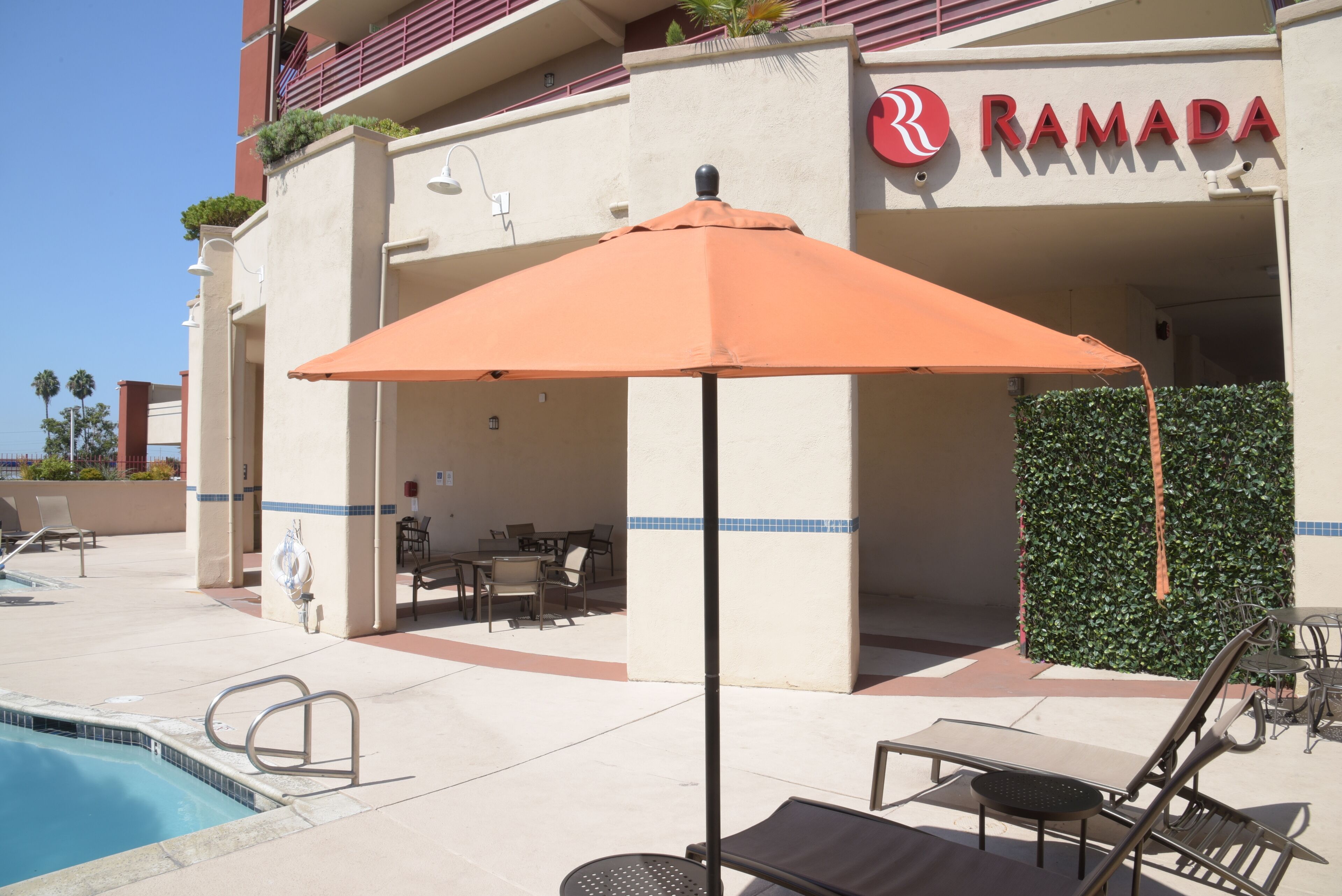 A poolside area with lounge chairs, an umbrella, and a sign for Ramada hotel.