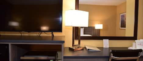 A hotel room with a flat-screen TV, a microwave, a desk with a lamp, a chair, and a large mirror.