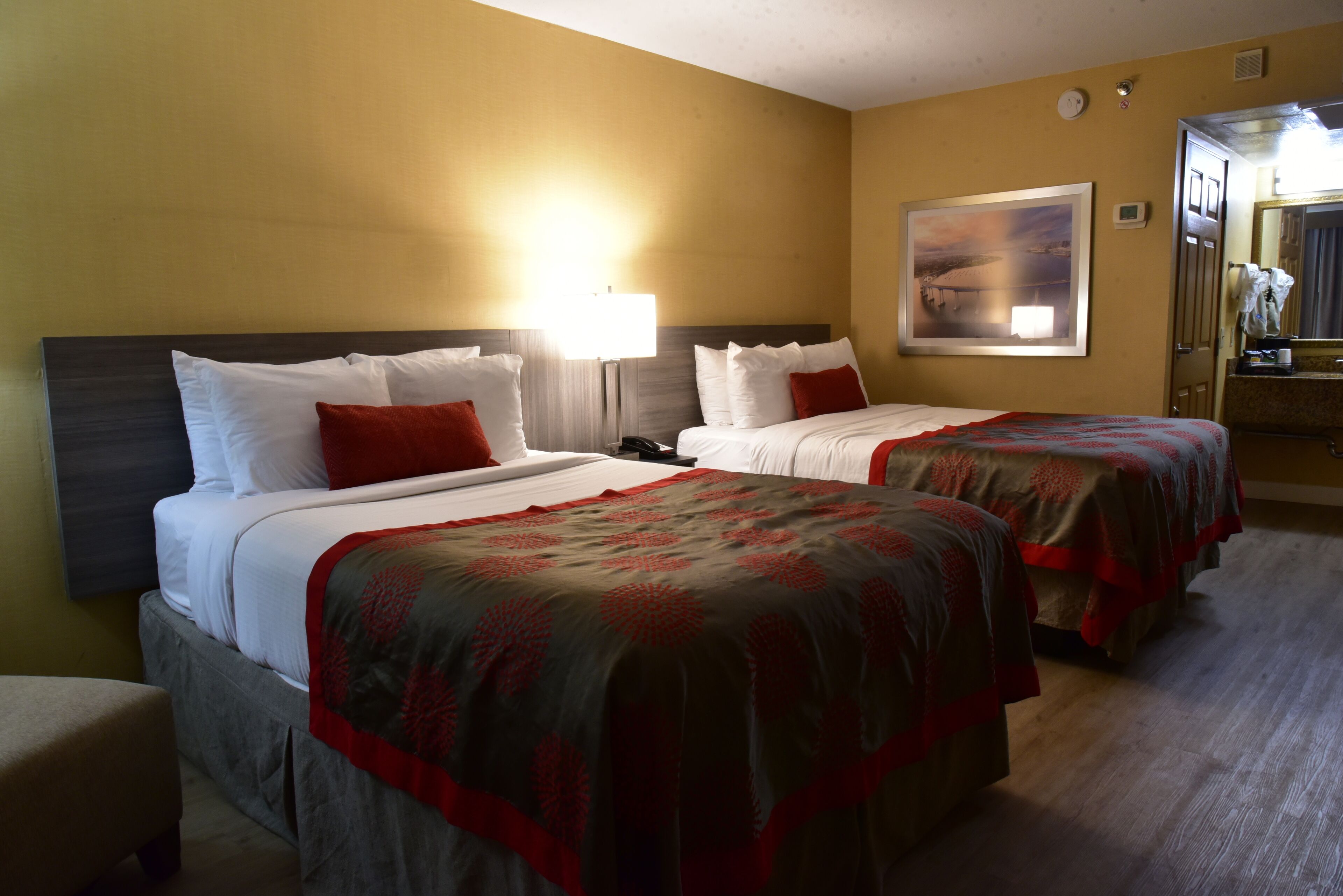 A hotel room with two beds, each with white linens and red pillows, a large painting on the wall, and a bedside table with a lamp.