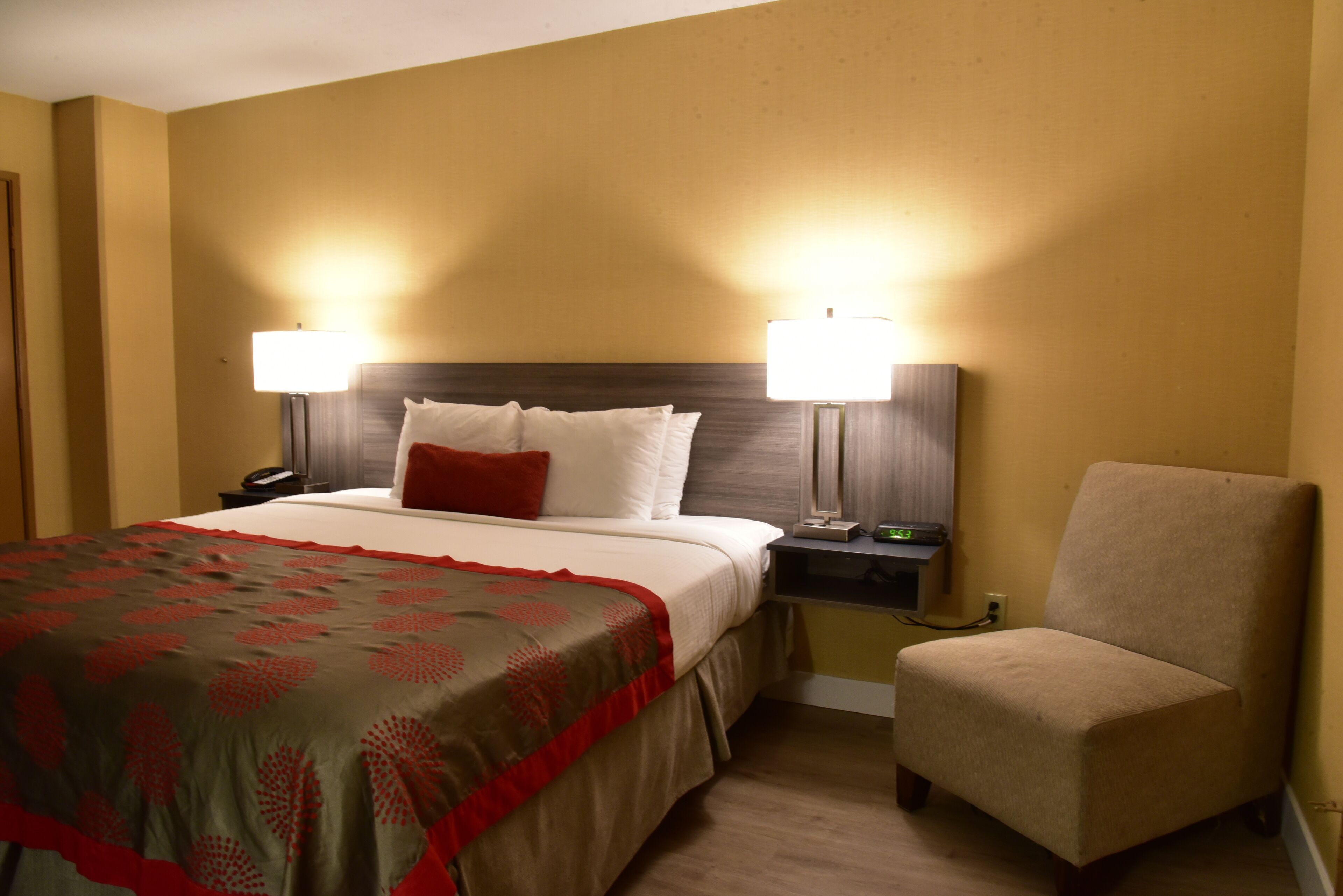 A hotel room with a bed, two bedside lamps, a chair, and a nightstand with a phone and a small table.