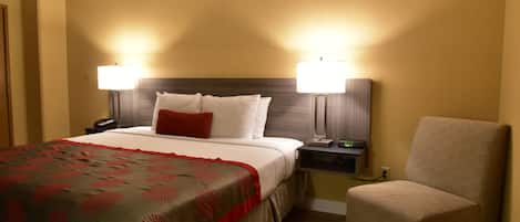 A hotel room with a bed, two bedside lamps, a chair, and a nightstand with a phone and a small table.