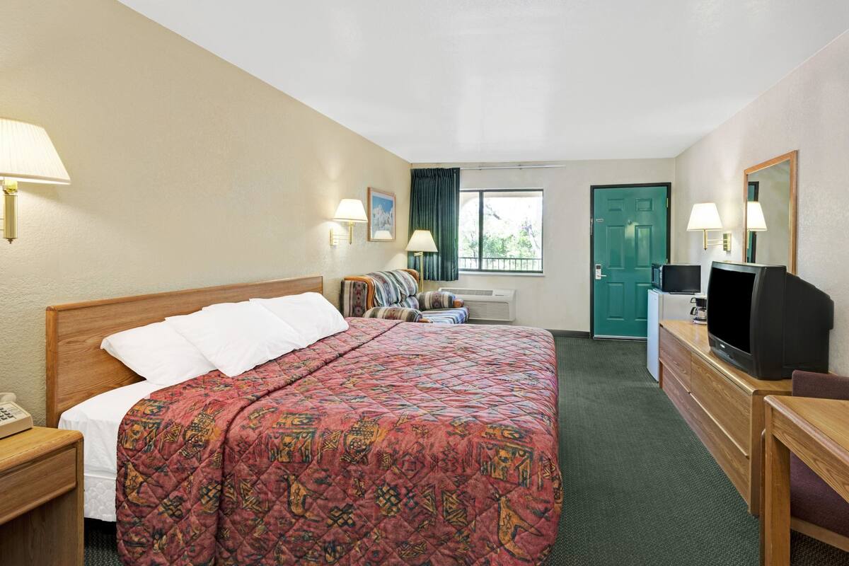A hotel room with a bed, a desk with a television, a chair, a desk lamp, a telephone, and a window with curtains.