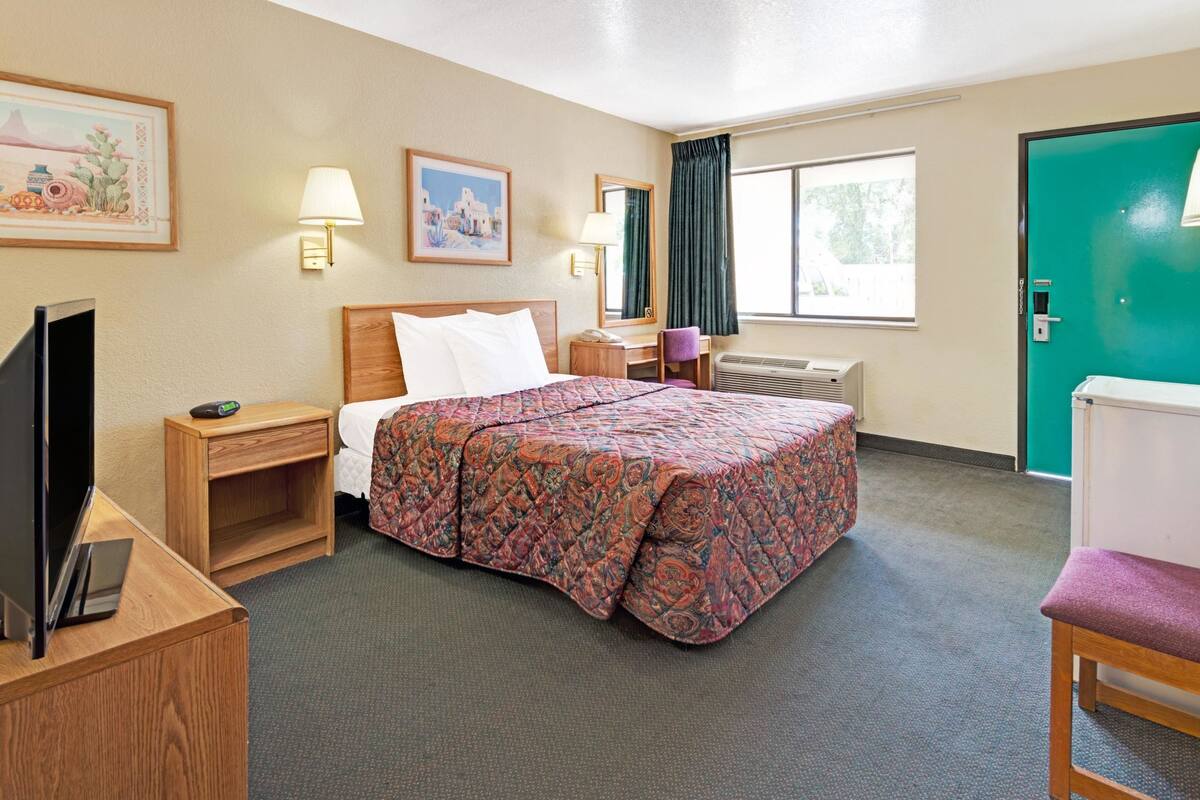 A hotel room with a bed, bedside table, lamp, desk, chair, and a television.