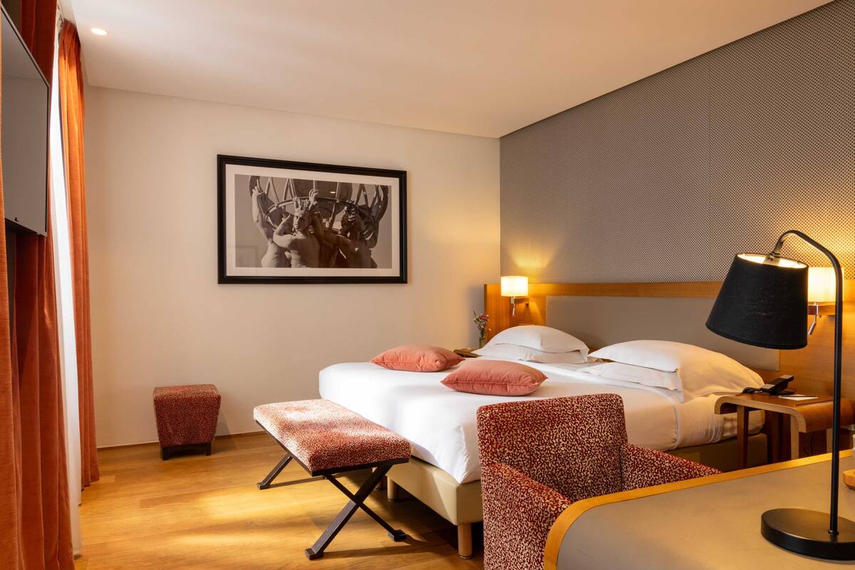 A hotel room with a bed, a bench, a chair, a nightstand, a lamp, and a framed picture on the wall.