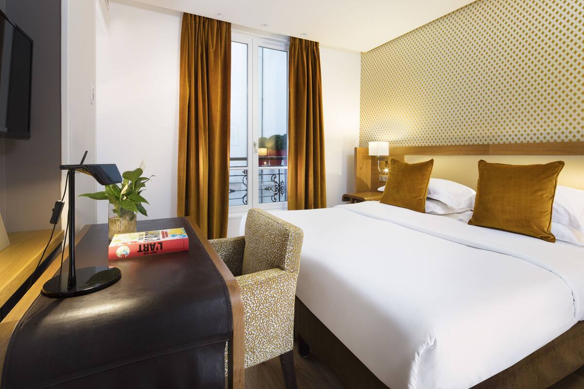 A hotel room with a bed, a desk with a lamp, a chair, a TV, and a window with curtains.