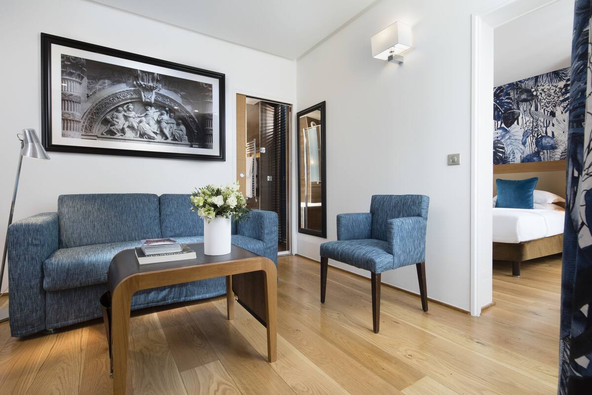 A modern living room with a sofa, armchair, and coffee table. There is a framed artwork on the wall and a door leading to another room.