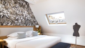 A hotel room with a large bed, a bench, a mannequin, a window with a city view, and a wall with a leaf pattern.