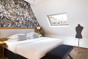 Habitación de hotel con una cama grande, un banco, un maniquí, una ventana con vista a la ciudad y una pared con un diseño de hojas.