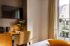 A hotel room with a bed, a desk with a TV, a chair, a vase with flowers, and a view of the outside.