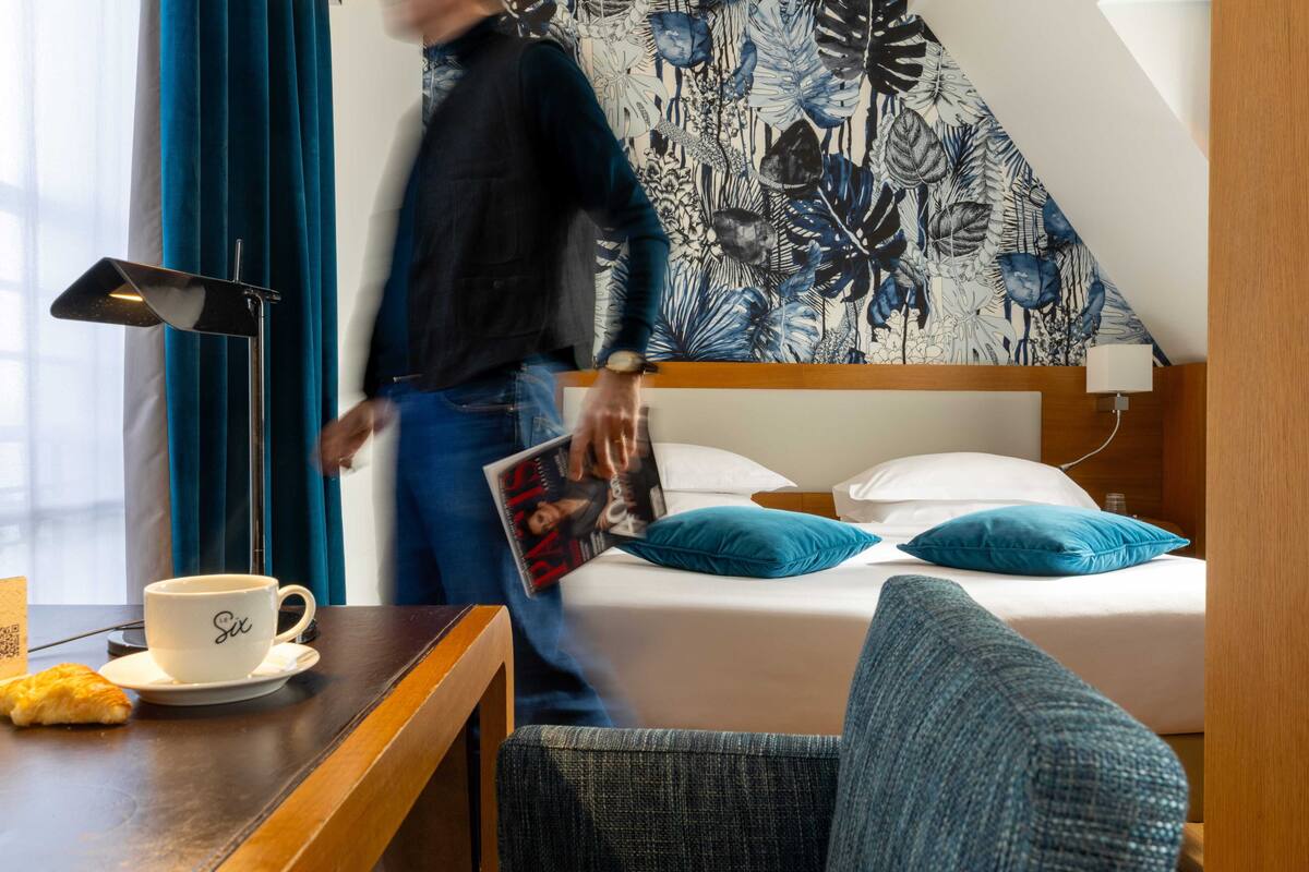 A hotel room with a bed, a bedside table with a cup and a pastry, a lamp, and a person walking by.