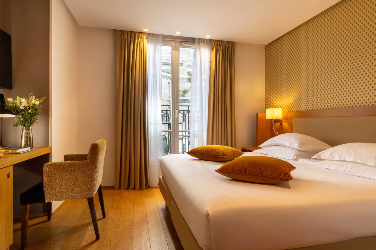 A hotel room with a bed, a desk, a chair, a TV, and a window with curtains.