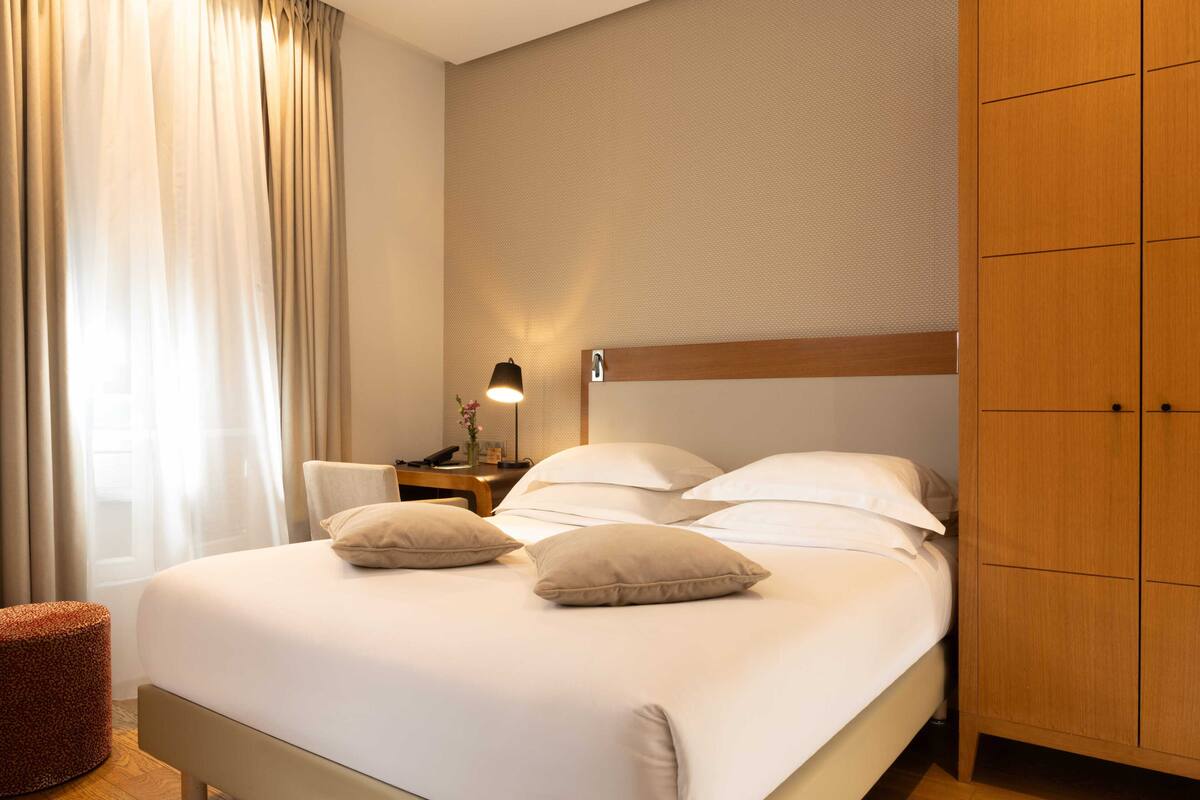 A hotel room with a bed, a nightstand, a lamp, a wardrobe, and a window with curtains.