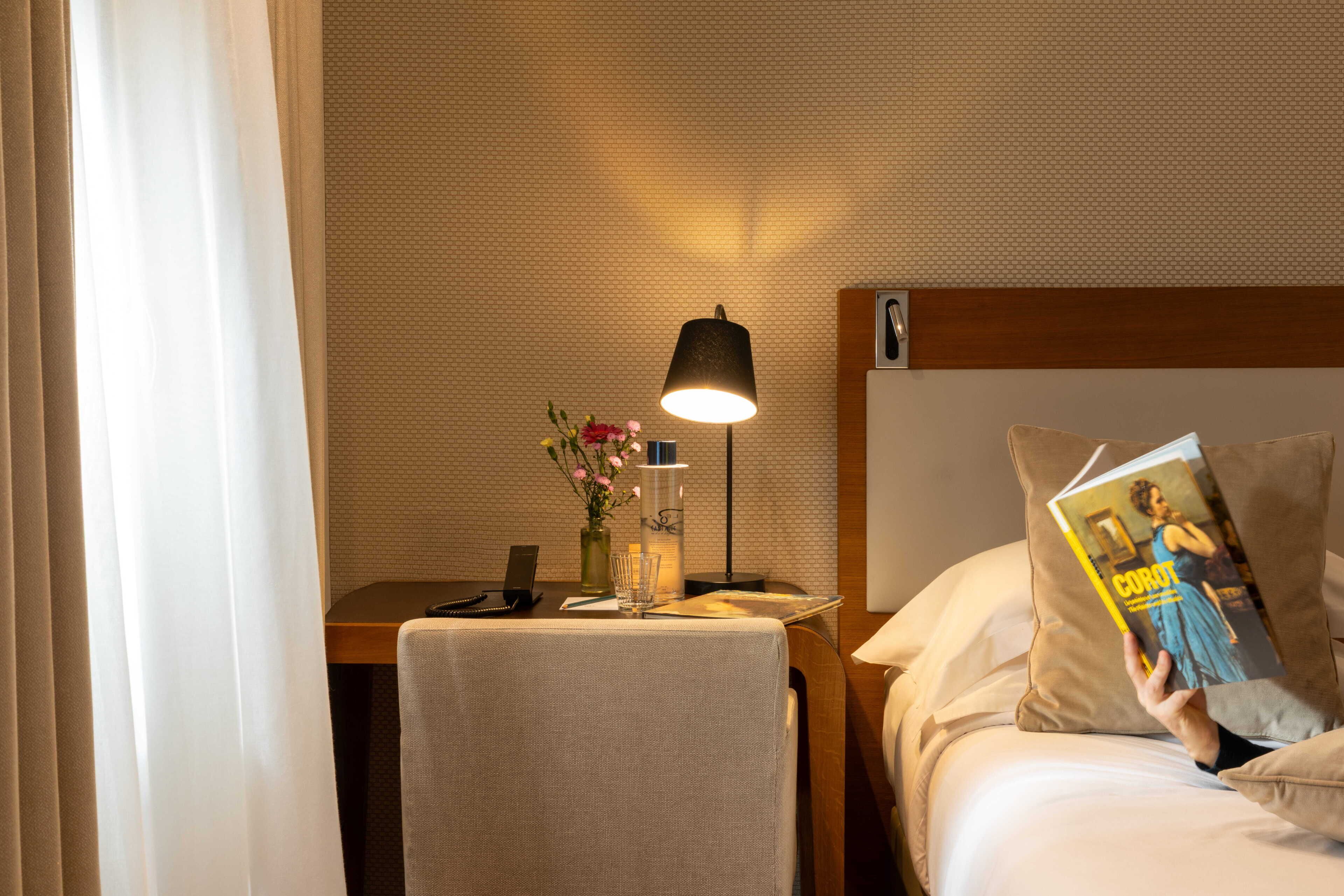 A hotel room with a bed, a bedside table with a lamp, a vase with flowers, and a book.